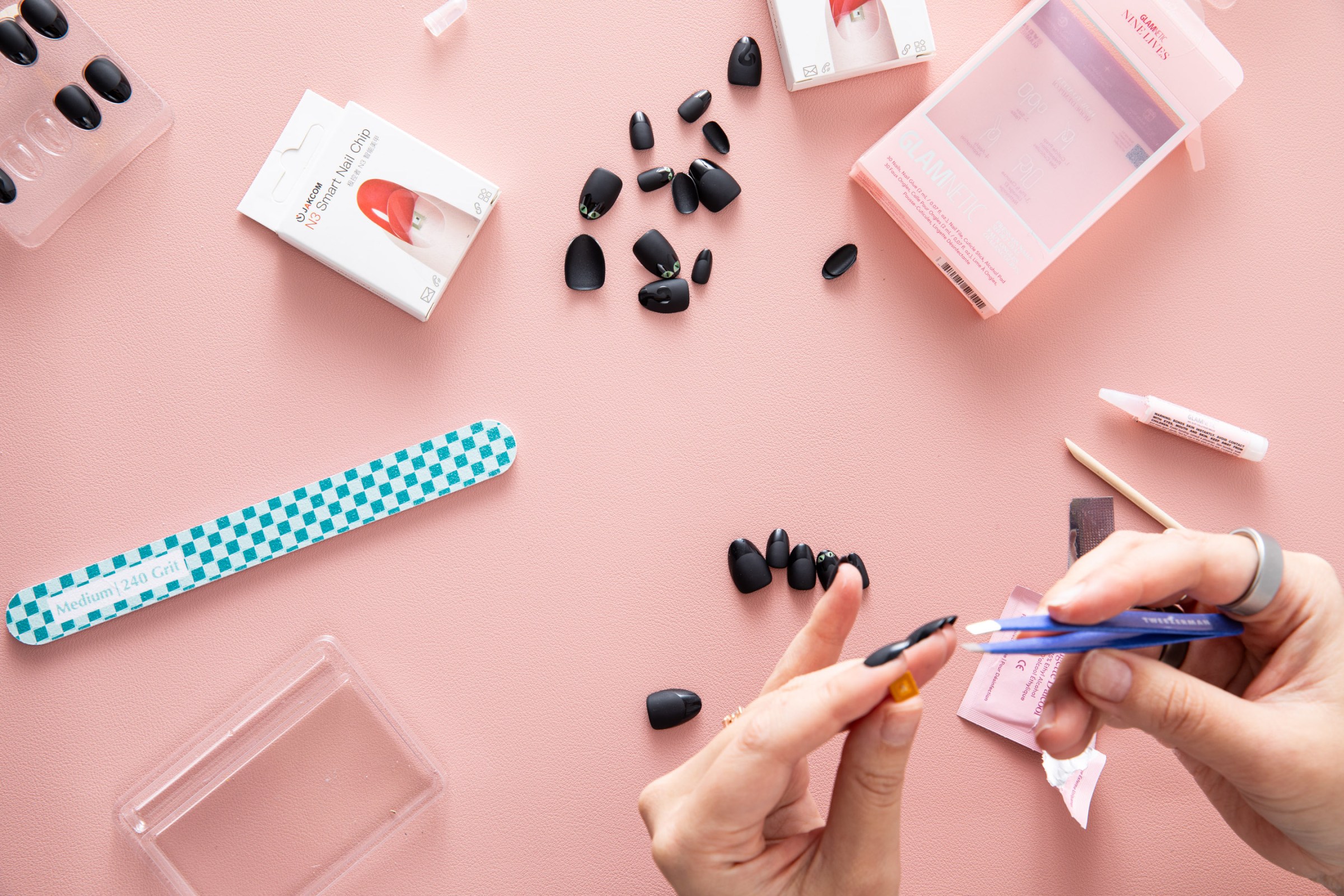 Top-down view of person applying Jakcom N3 Smart Nail chip to their nail. The supplies are scattered about, including a nail file, lots of matte black nails, empty boxes, cuticle stick, alcohol pad, and nail glue.