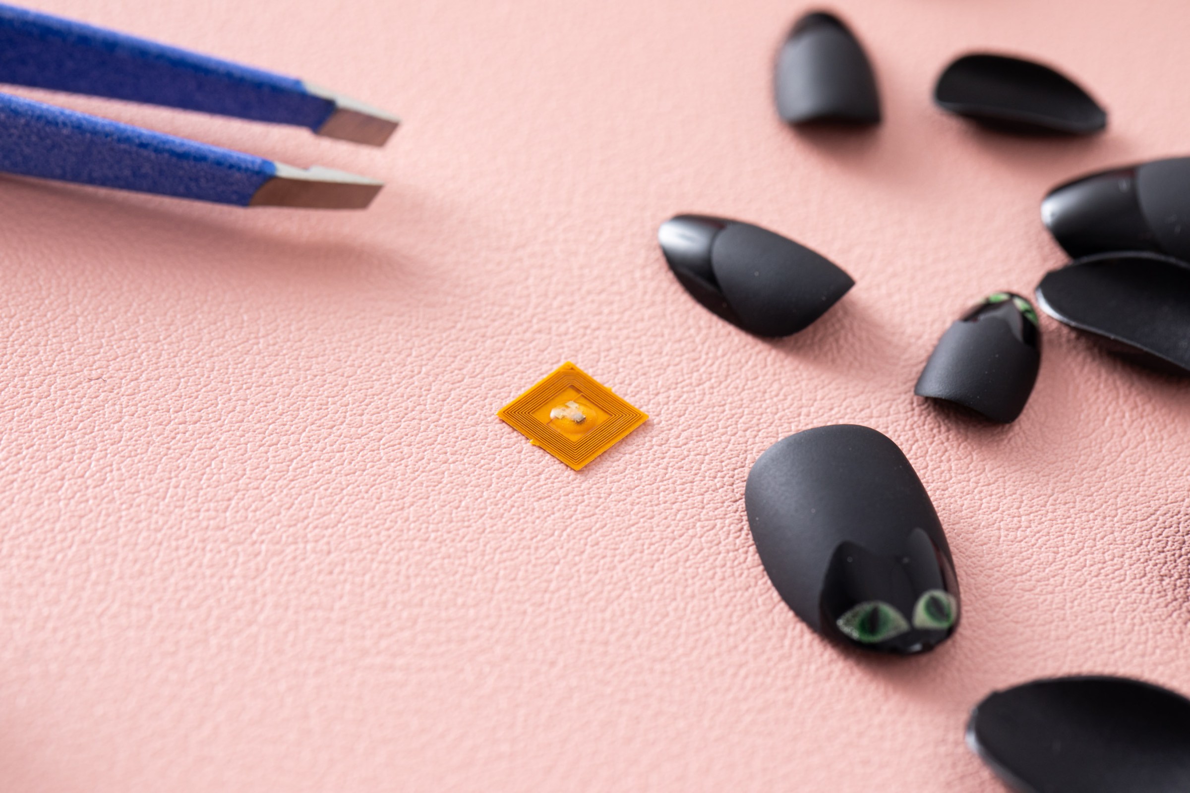 Closeup of the Jakcom N3 Smart Nail Chip next to black matte press on nails with a cat face. Tweezers are in the background. Everything is on a pink mat.
