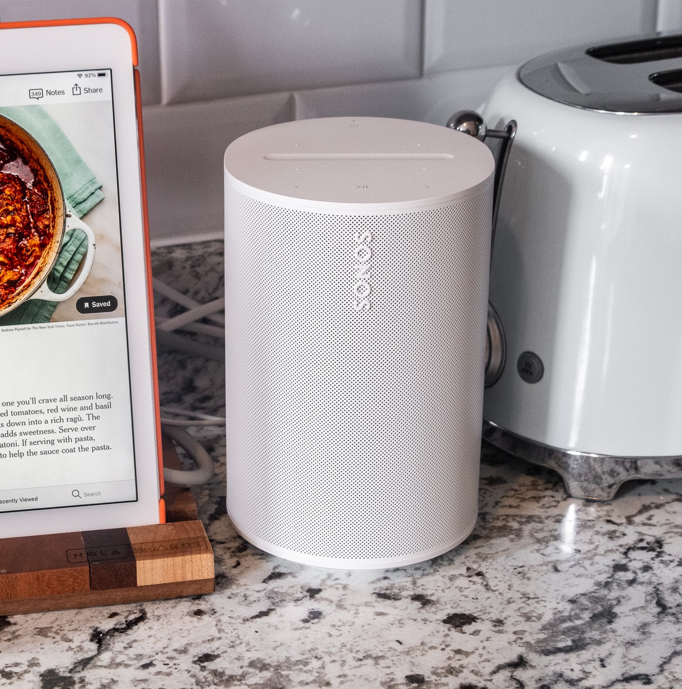 A photo of the Sonos Era 100 speaker in a kitchen setting beside an iPad and toaster.