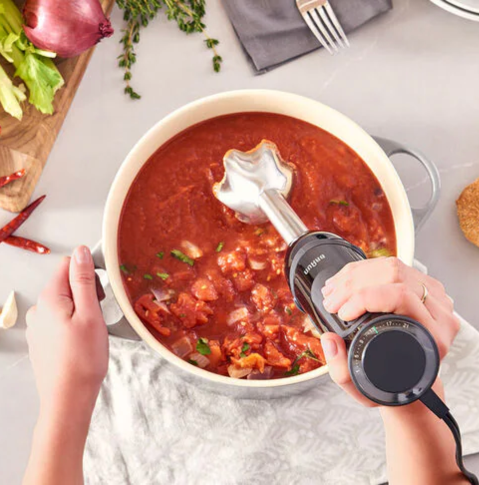 Overhead shot of hands holding a hand blender and stirring a bowl of a tomato-y broth.