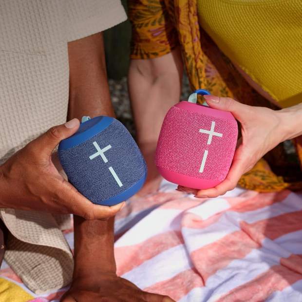 Two people holding UE WonderBoom 4 speakers, one blue and one pink.