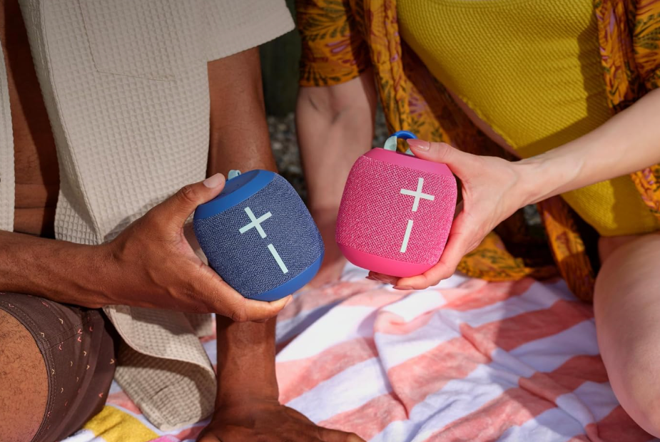 Two people holding UE WonderBoom 4 speakers, one blue and one pink.
